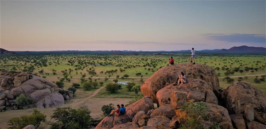Pessoas sentadas em cima de pedras grandes, apreciando a paisagem de Damaraland, na Namíbia, com bastante árvores, gramado verde e horizonte montanhoso