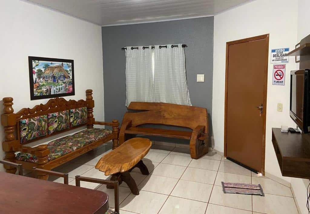 Sala de estar com sofá de madeira, banco, mesa de centro, quatro decorativo, janela com cortina, TV em painel, mesa, tapetinho e avisos de "proibido fumar" e de "desligar a luz e o ar-condicionado"