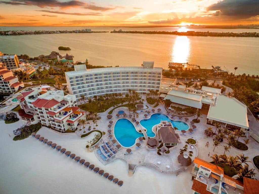 vista área do sol se pondo no mar atrás do prédio Grand Park Royal Cancún resort, onde é possível enxergar as piscinas e espreguiçadeiras do hotel