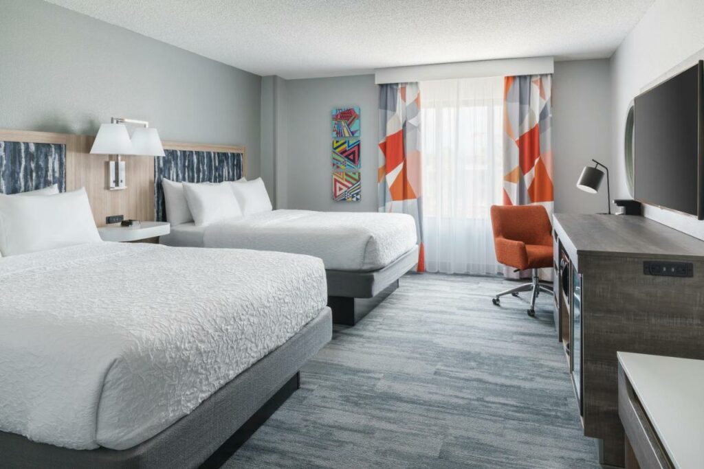 Quarto do Hampton Inn & Suites Tampa Ybor City Downtown com chão de madeira acinzentada, uma cama de casal e uma de solteiro, uma mesa de trabalho e uma cadeira estilo poltrona, uma janela com cortinas, uma televisão e uma cômoda, tudo em tons de laranja e cinza