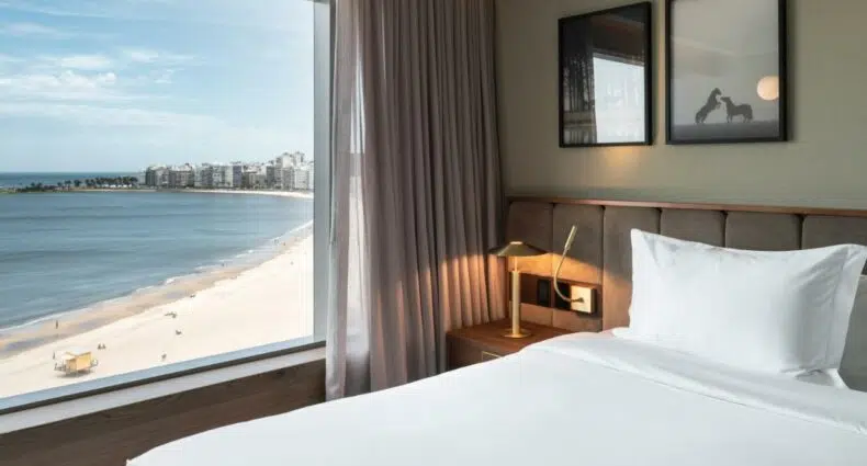 Quarto do Hotel Costanero Montevideo- MGallery, com uma cama feita, uma luminária de mesa e uma grande janela com vista para a praia e o oceano, para representar o post de hotéis em Montevidéu