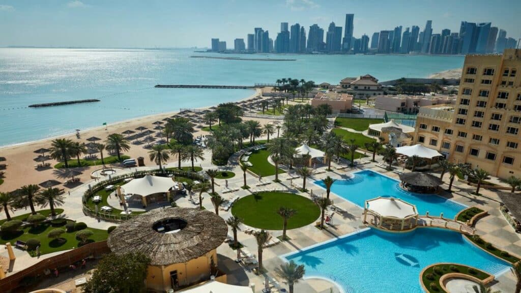 Visão aérea do hotel InterContinental Doha Beach & Spa, uma das recomendações do post seguro viagem Doha. Há duas piscinas na frente do hotel e algumas áres verdes. Ao fundo, podemos ver prédios e o mar turquesa.