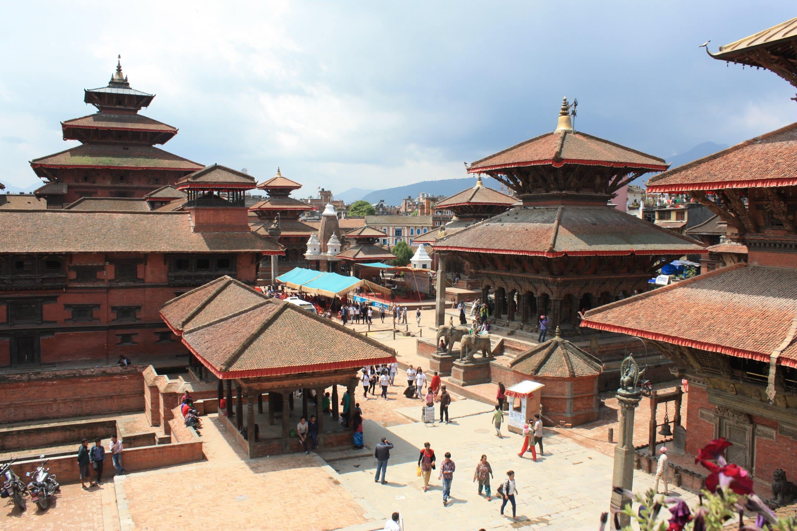 Templos marrom e vermelhos embaixo do céu azul para ilustrar o post de seguro viagem Nepal. Há pessoas caminhando entre as construções. - Foto: via Pxhere