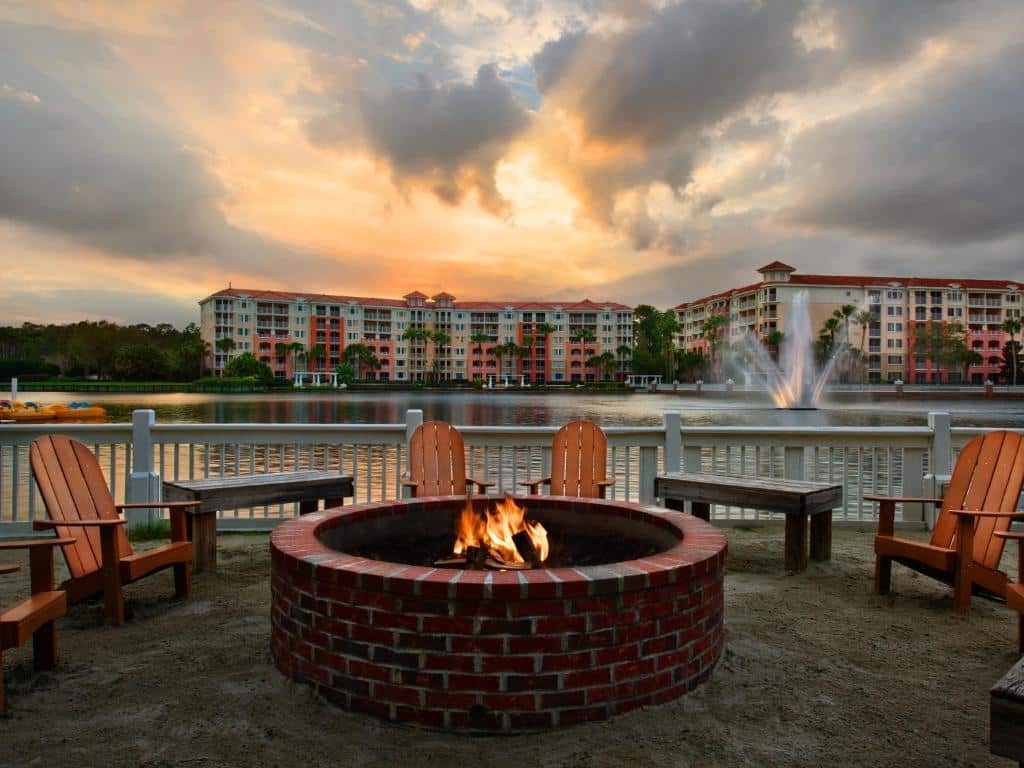 cadeiras com uma espécie de lareira redonda no meio pegando fogo, com vista para o lago e construções do Marriott's Grande Vista atrás, sendo uma das opções de hotéis em Orlando