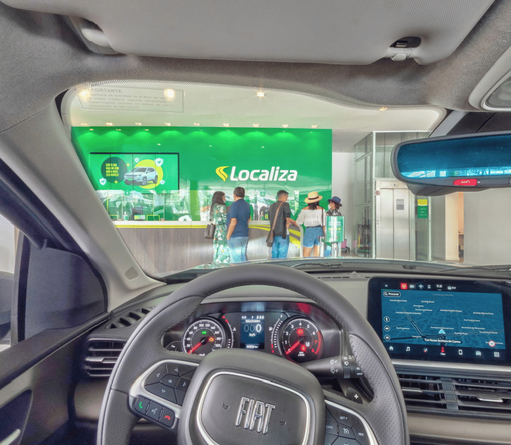 Foto de dentro de um carro Fiat Pulse parado em frente a um balcão da Localiza Rent a Car, com cinco pessoas esperando atendimento em frente