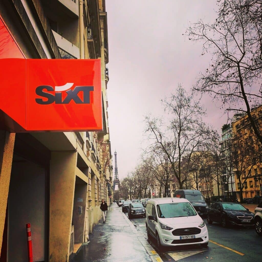 Placa de loja da Sixt em rua de Paris, com a Torre Eiffel ao fundo, prédios clássicos parisienses em ambos os lados da rua, uma pessoa andando na calçada e carros estacionados ao longo da rua