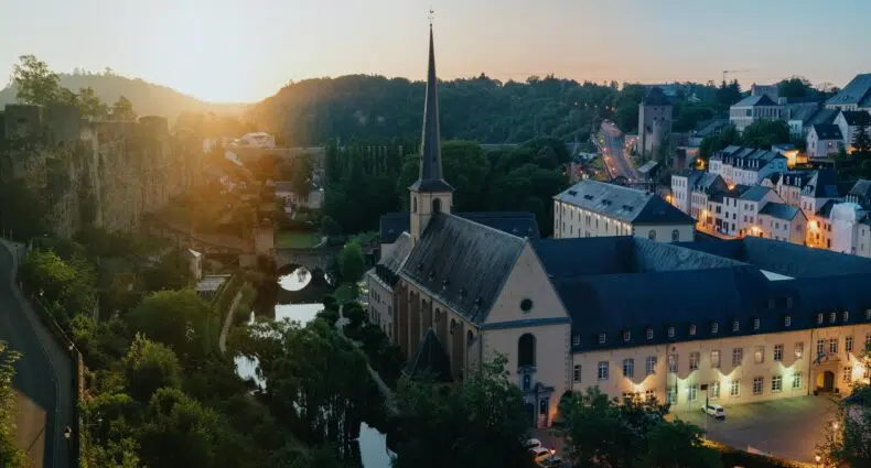 neumünster abbey com árvores ao lado e algumas casas com o sol iluminando ilustrando post de seguro viagem Luxemburgo