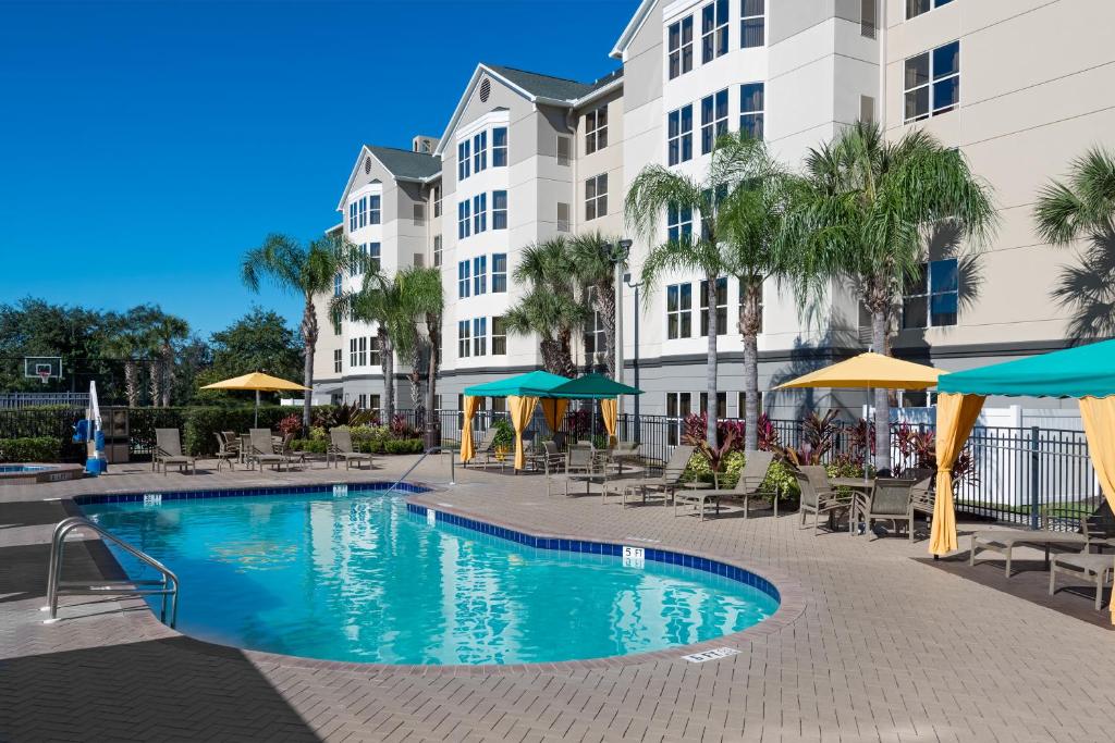 piscina com bordas arredondadas, com vários guarda-sóis quadrados azuis e amarelos com o prédio do Homewood Suites by Hilton Orlando-Nearest to Universal Studios atrás, sendo um dos hotéis em Orlando