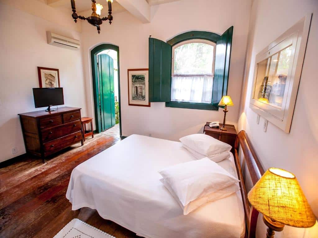 Quarto na Pousada da Marquesa com uma cama de casal, uma cômoda com uma televisão, duas mesinhas de cabeceira com abajures, uma janela e um ventilador de teto, o estilo é colonial, com tudo em madeira e alguns detalhes em verde