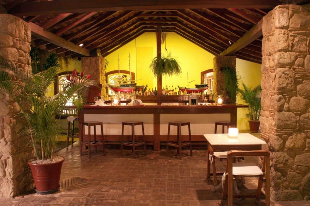 Restaurante rústico e com paredes de pedra na Pousada do Ouro, com mesinhas e cadeiras de madeira e um bar em destaque no centro da foto