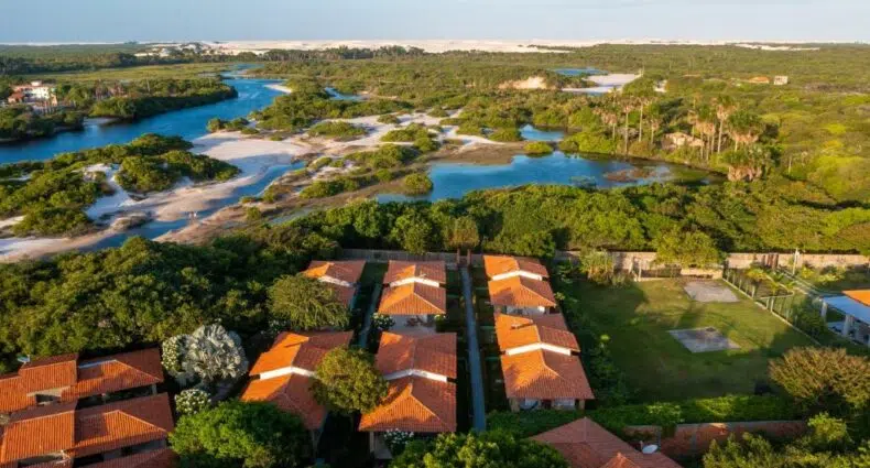 Vista aérea de casas com telhado laranja cercadas por vegetação, perto de um rio e de uma paisagem arenosa sob um céu claro.