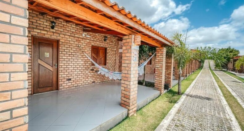 Casa de tijolos com telhado de telha, rede na varanda e um caminho de paralelepípedos ao lado de palmeiras sob um céu azul.