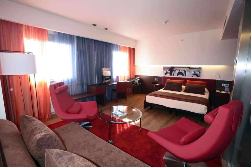 quarto com decoração vermelha com cama de casal, poltronas e mesa de vidro, duas janelas grandes com cortina e sofá do Ayre Gran Hotel Colón em Madri