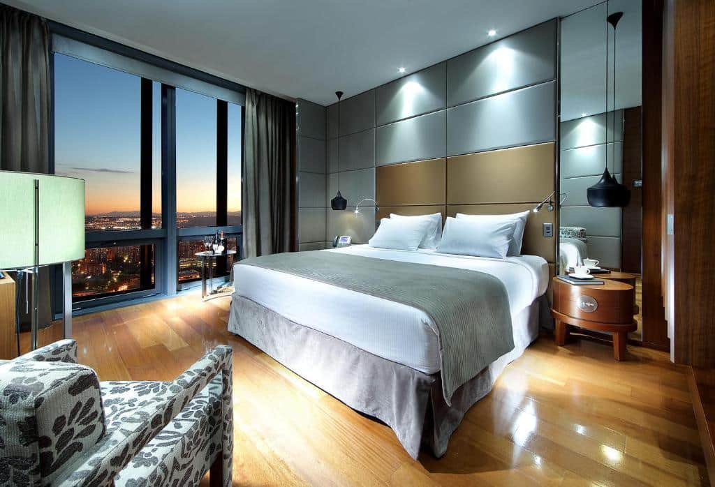 quarto com cama e janela enorme, que pega a parede inteira, com vista da cidade e poltrona e tv no Eurostars Madrid Tower