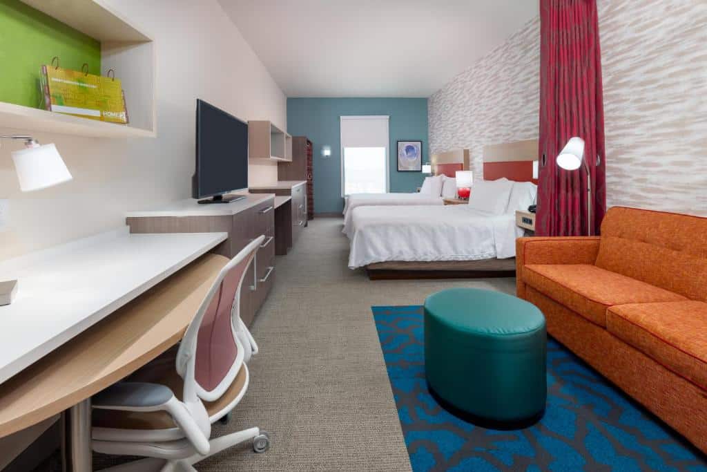 quarto amplo com duas camas, mesa de trabalho, sofá e puff, tudo em tons coloridos de laranja e azul no Home2 Suites By Hilton Orlando Near Universal
