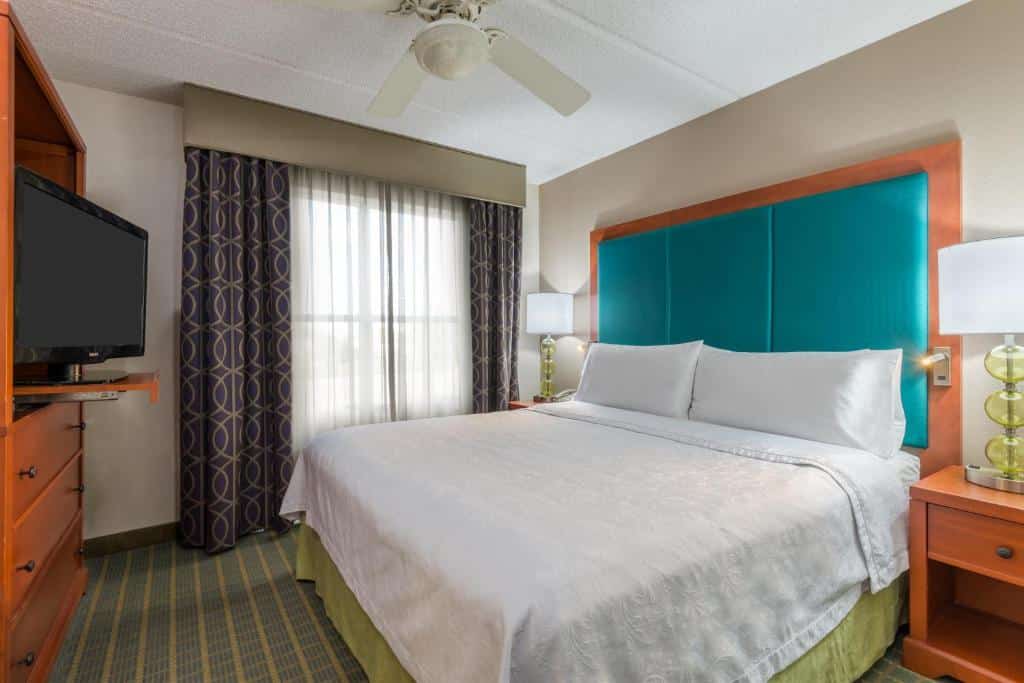 quarto com cama de casal, janela grande e cabeceira azul, com tv e móveis de madeira no Homewood Suites by Hilton Orlando-Nearest to Universal Studios