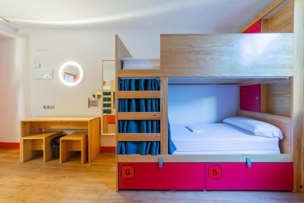 Quarto do Ok Hostel Madrid, uma das recomendações de hostels em Madri. O beliche é bem estruturado e tem uma cortina de privacidade na cama debaixo. Do lado esquerdo da foto, uma mesa de madeira com dois bancos tem um espelho na parede acima.