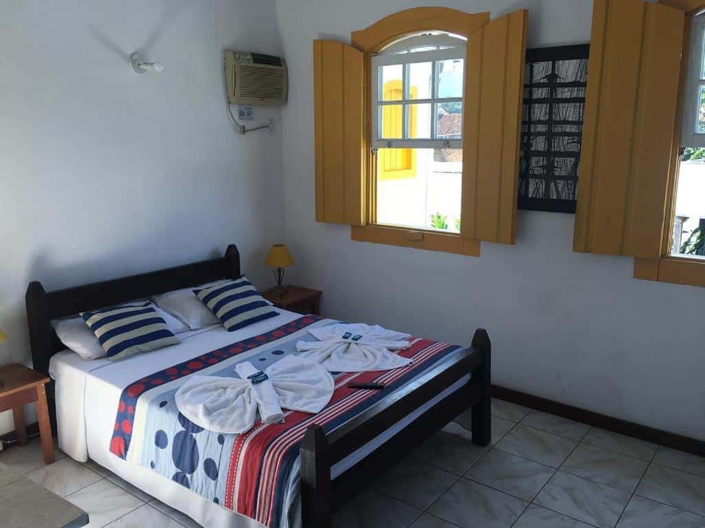 quarto com cama de casal com colcha colorida e janelas amarelas na Pousada Fortaleza
