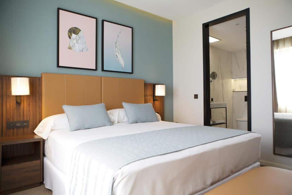 Quarto do Riu Plaza España, uma das recomendações de hotéis de luxo em Madri. A cama grande tem jogo de cama branco e azul, e a cabeceira é acolchoada no centro e de madeira nas bordas, com mesinha e abajur. A porta do banheiro fica ao lado e está aberta, há também um espelho ao lado da porta. A parede da cama é azul e possui dois quadrinhos.