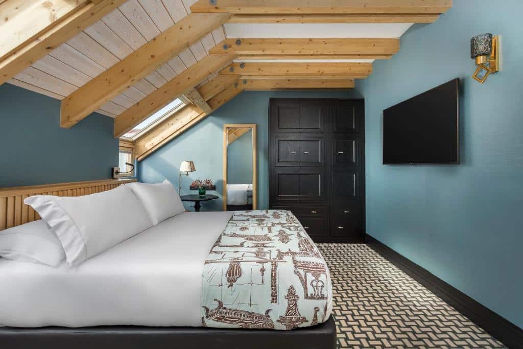 quarto com cama grande, parede azul com teto aparecendo as vigas, ou seja, com detalhes em madeira, guarda-roupa e espelho no Room Mate Alba