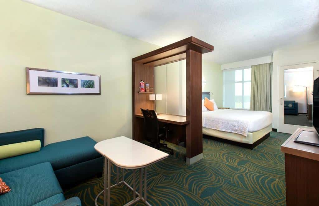 quarto grande com uma separação de vidro em que é uma escrivaninha, de um lado um sofá azul com mesinha e do outro a cama de casal com tv do SpringHill Suites by Marriott Orlando at FLAMINGO CROSSINGS Town Center-Western Entrance, um dos hotéis em Orlando