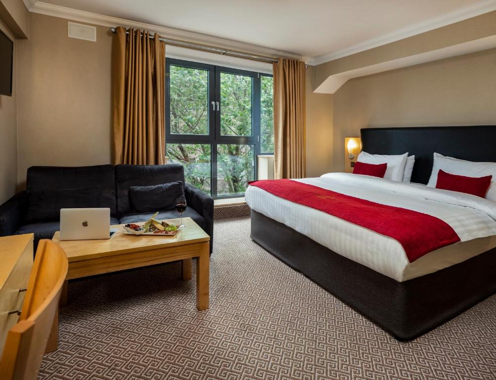 Quarto espaçoso no Academy Plaza Hotel com uma janela ampla, uma sofá, uma mesa de centro, uma cômoda e uma cama de casal, tudo em tons mais neutros, apenas a roupa de cama é vermelha