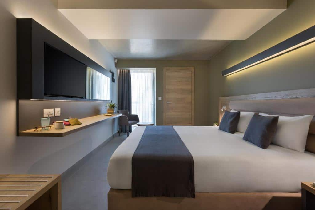 Quarto do Azur Hotel by ST Hotels com uma cama de casal, uma televisão, uma pequena mesinha e uma poltrona, ambiente clean, em tons de branco e cinza