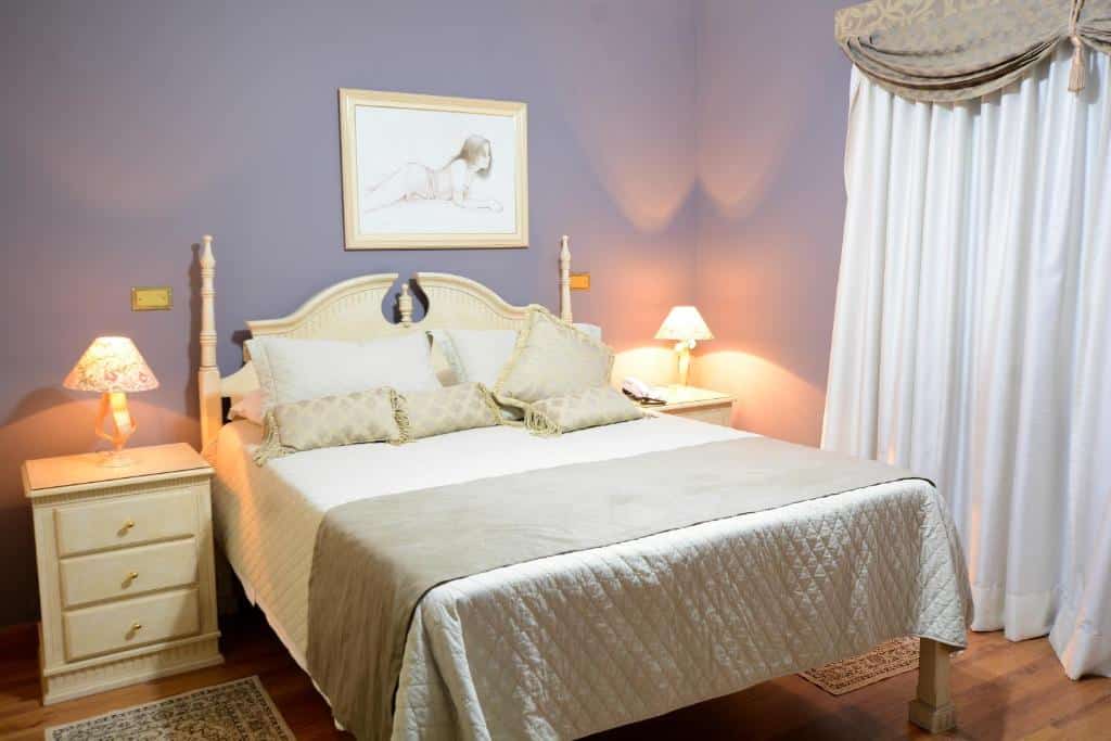 Quarto da Biazi Paradise Hotel com uma cama de casal, duas pequenas cômodas com abajures sob elas, as paredes têm um tom de lilás e uma janela com cortinas