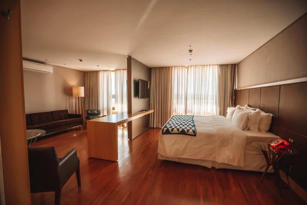 Quarto amplo no Brasília Palace Hotel com uma cama de casal, uma varanda, um sofá e uma poltrona, uma televisão e uma bancada que divide os dois ambientes, o chão é de madeira e toda a decoração é em tons de marrom