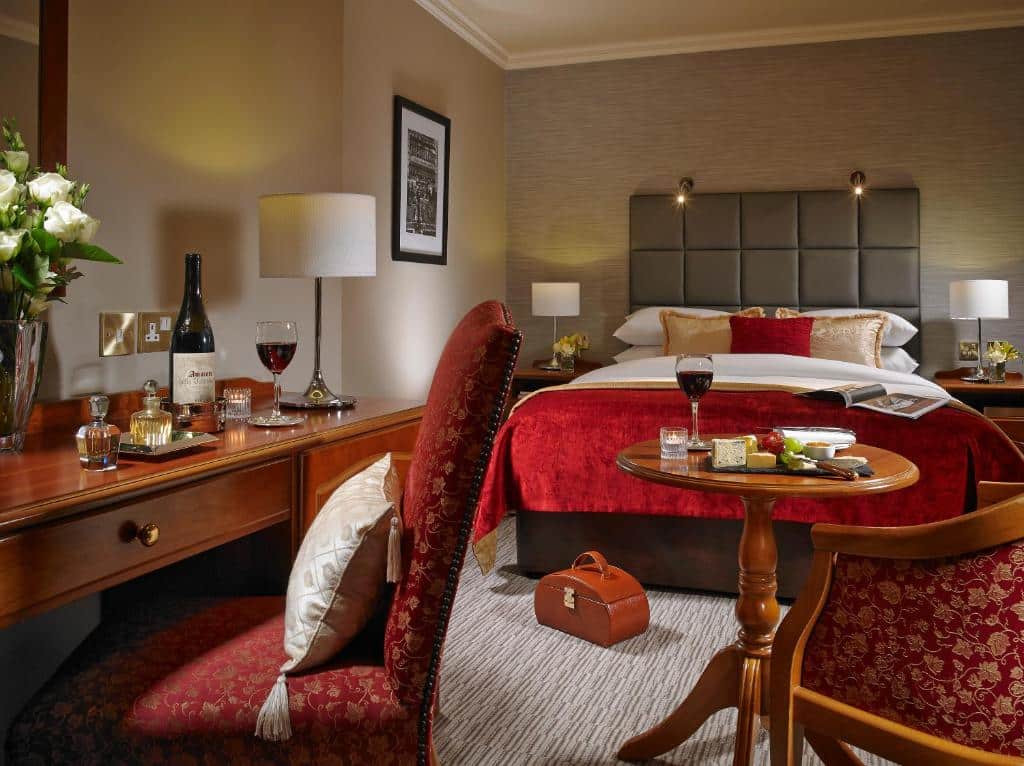 Quarto no Buswells Hotel com uma cama de casal, uma pequena mesa de centro com uma poltrona, uma mesa de escritório com uma cadeira, alguns itens de coração, duas mesinhas de cabeceira ao lado da cama com abajures, tudo decorado em tons de vermelho e dourado