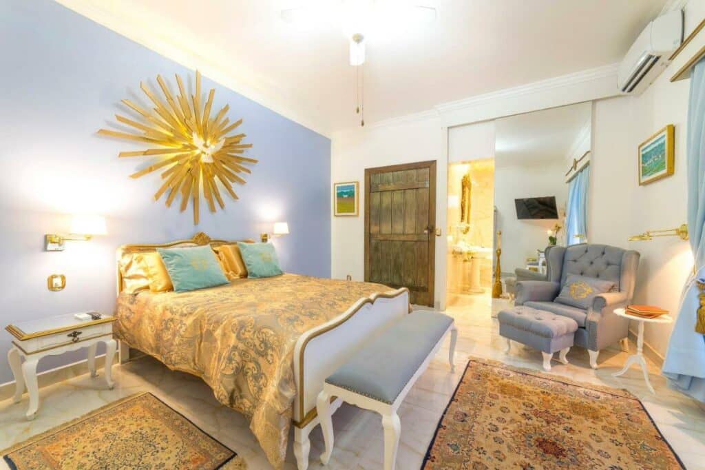 Quarto amplo na Casa Nostra Boutique Hotel com uma decoração bem colonial em tons de dourado, azul e branco, com uma cama de casal, uma poltrona, um ar-condicionado, uma janela ampla, e outros itens de decoração