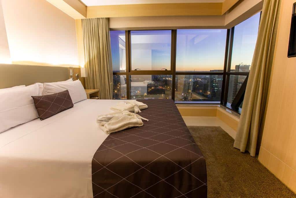 Quarto no Cullinan Hplus Premium com uma janela ampla dando vista para a cidade, uma cama de casal com travesseiros e roupões sobre a cama, para representar hotéis em Brasília