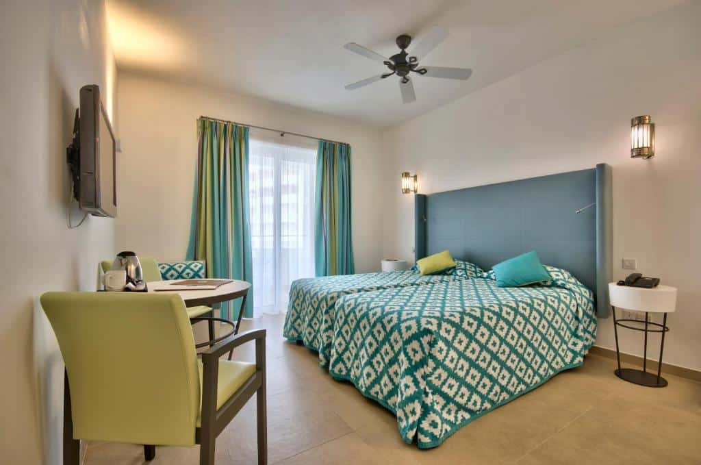Quarto no db San Antonio Hotel + Spa All Inclusive com duas camas de solteiro, uma varanda, um ventilador de teto, uma mesinha com duas cadeiras e uma televisão, tudo decorado em tons de branco, verde claro e azul claro