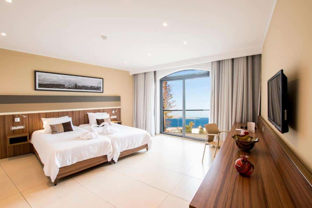 Quarto amplo no Dolmen Hotel Malta com uma sacada dando vista para o mar, uma televisão, duas camas de solteiro e uma cabeceira com duas luminárias