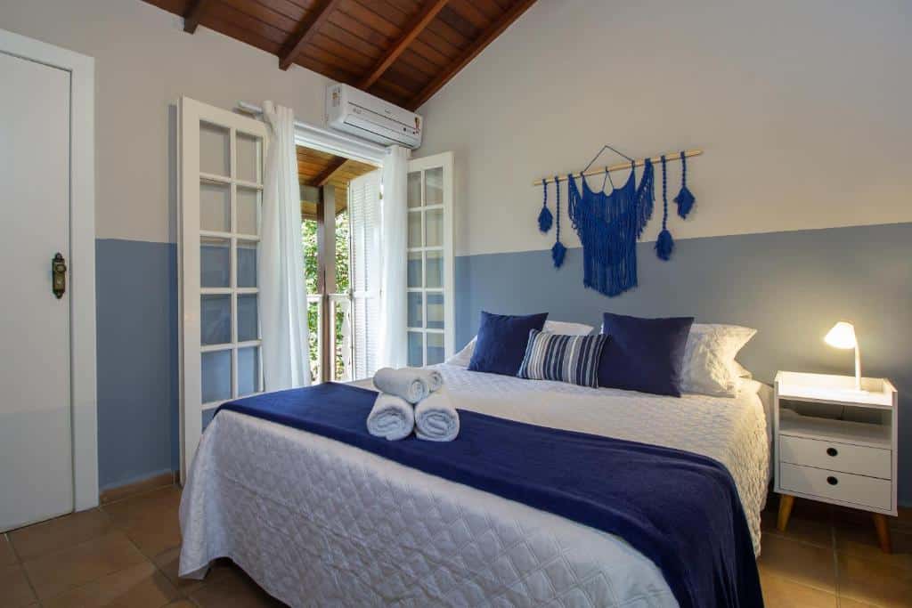 Quarto da Estrela do Mar Paraty com tudo decorado em branco e azul, a cama de casal é coberta por toalhas, travesseiros e almofadas, há uma sacada e um banheiro, assim como um pequeno móvel com um abajur do lado da cama