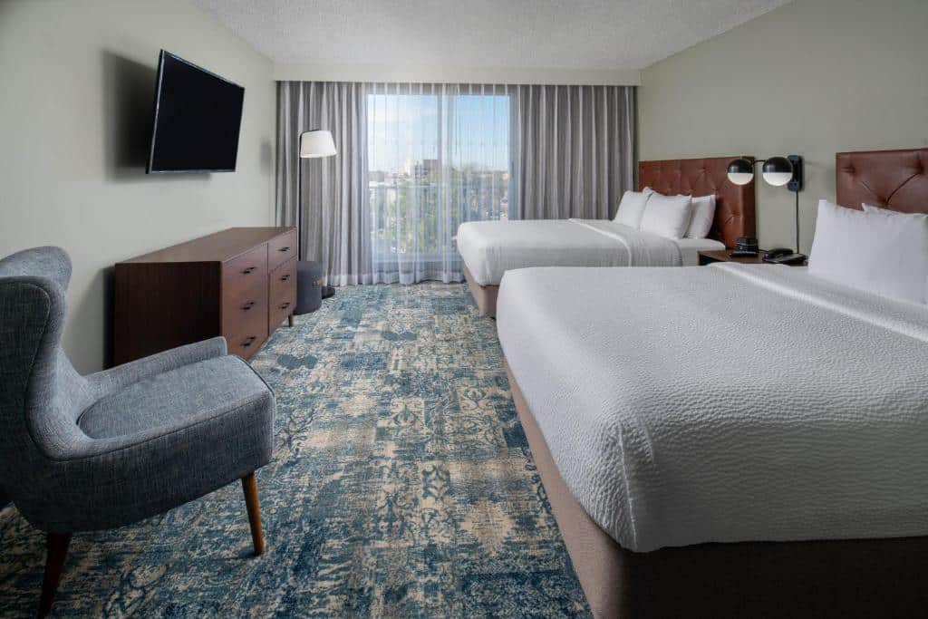Quarto no Four Points by Sheraton Suites Tampa Airport Westshore com uma cama de casal, uma de solteiro, uma ampla sacada, uma cômoda, uma poltrona e uma televisão, tudo em tons de branco, cinza e azul