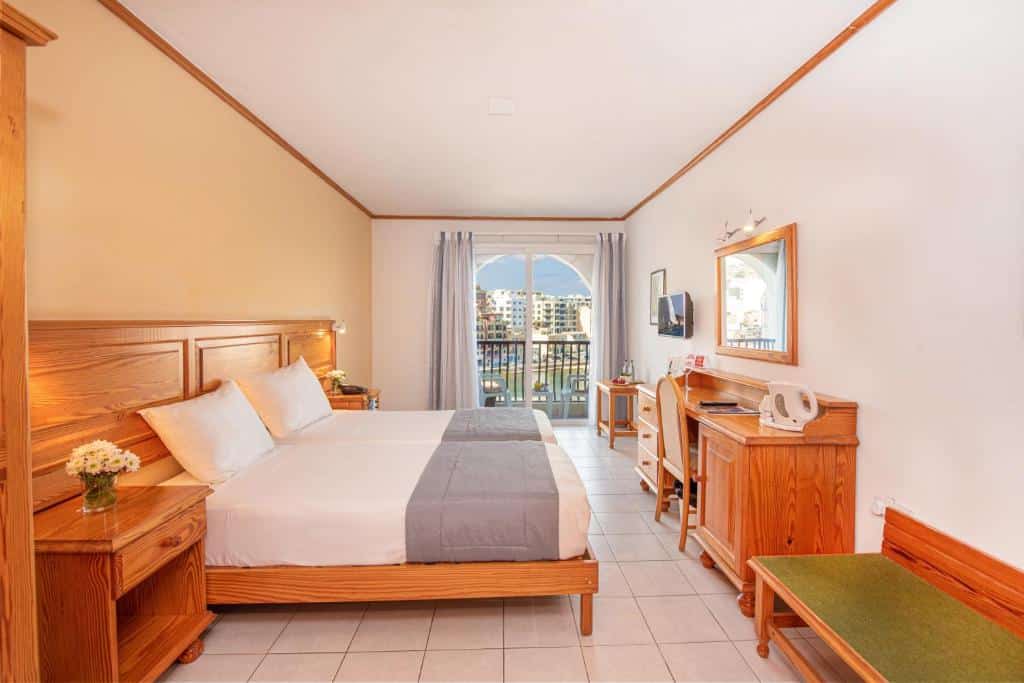 Quarto no Gillieru Harbour Hotel com uma cama de casal, uma varanda com cortinas, uma cômoda com uma cadeira, um espelho, tudo decorado em tom de madeira clara e roupas de cama brancas