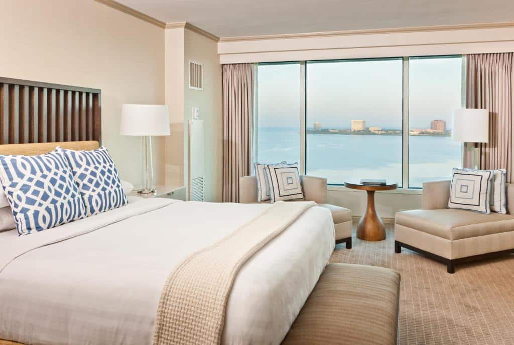 Quarto do Grand Hyatt Tampa Bay com uma cama de casal, uma mesinha com duas poltronas perto de uma janela ampla com vista para o mar, chão de carpete bege, toda a decoração está em tons de bege claro e branco
