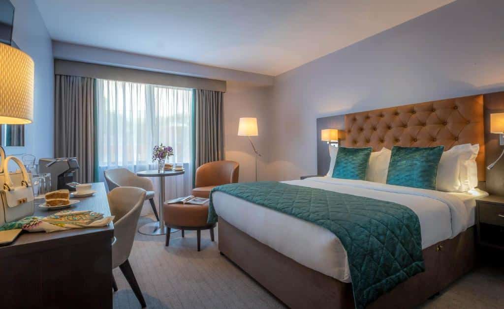 Quarto do Grand Canal Hotel com uma cama de casal, uma janela, duas poltronas e uma pequena mesinha, uma mesa de escritório com uma cadeira, um espelho e dois abajures de chão, para representar os hotéis em Dublin