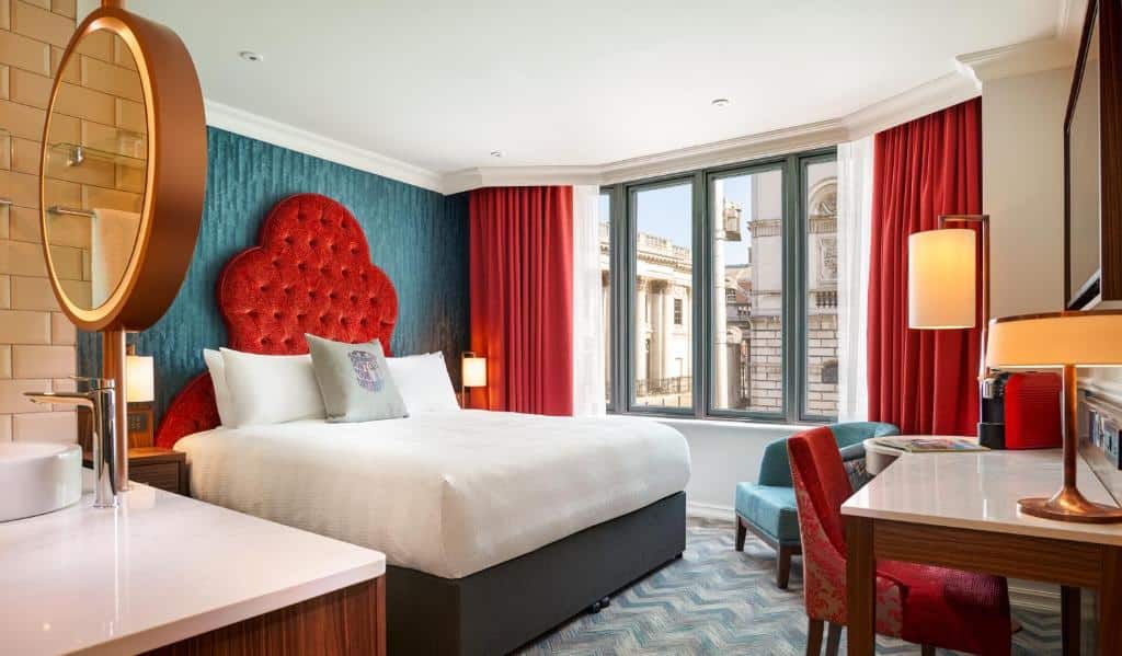 Quarto no Hard Rock Hotel Dublin com uma janela ampla com cortinas vermelhas, uma poltrona, uma mesa de trabalho com uma cadeira, uma televisão, uma cama de casal, tudo decorado em tons de azul turquesa, vermelho e branco