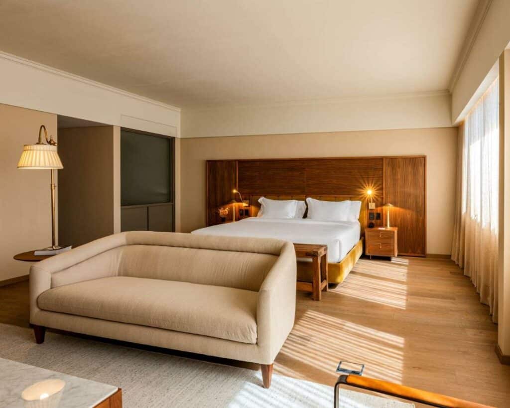 Quarto amplo no Hotel Costanero Montevideo- MGallery com tudo em madeira, janela com cortinas, uma cama de casal, uma sofá bege, uma cabeceira com dois pequenos móveis com abajures nas laterais da cama, para representar hotéis em Montevidéu