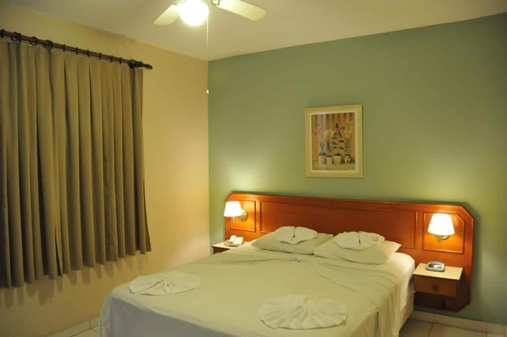 Quarto do Hotel Fazenda São Matheus com uma cama de casal, uma cabeceira de madeira com abajures instalados, uma janela com cortinas, tudo em tons de verde, para representar hotéis em Serra Negra