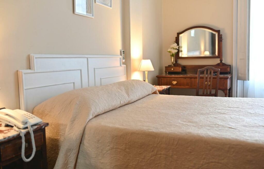 Quarto no Hotel Palacio com um estilo mais vintage, com uma cama de casal, uma penteadeira, um espelho, uma cadeira, um abajur, tudo em tons de madeira e as paredes brancas, para representar hotéis em Montevidéu