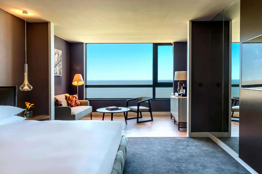 Quarto no Hyatt Centric Montevideo com uma cama de casal, uma pequena ante sala com vista para o mar, com um sofá, uma mesinha e uma cadeira, um abajur de chão e uma televisão, para representar os hotéis em Montevidéu