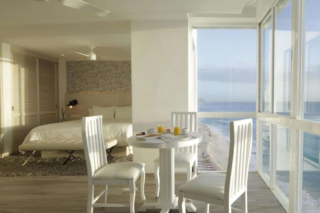 suíte do Oleo Cancun Playa Boutique com uma cama de casal, uma mesa redonda com três cadeiras e um café da manhã preparado em cima, com vista para a praia pelas janelas amplas do quarto