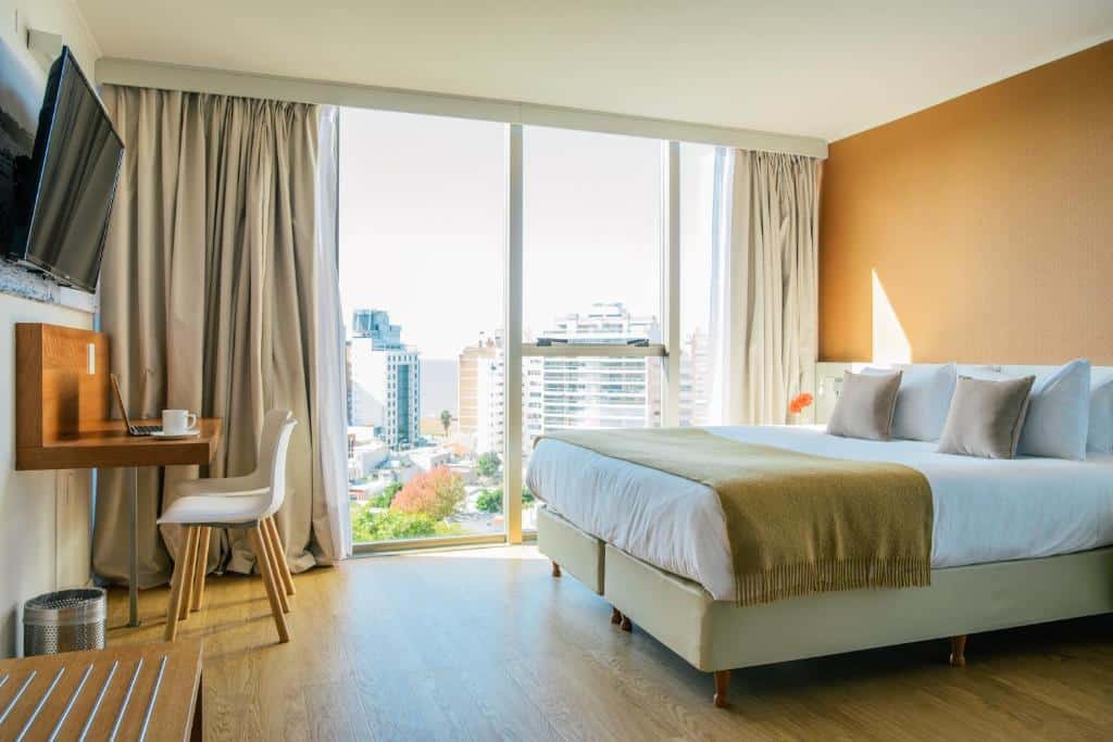 Quarto no Own Montevideo com cama de casal, piso de madeira, uma janela ampla, uma mesinha de escritório com duas cadeiras e uma televisão