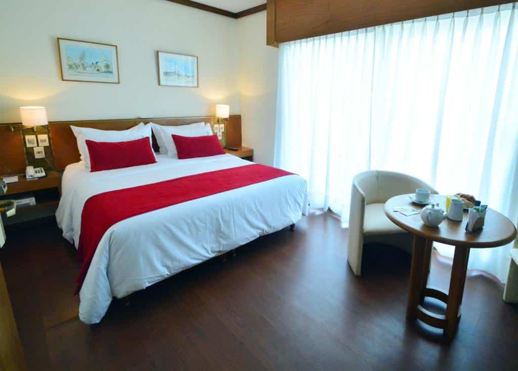 Quarto no Oxford Hotel com piso de madeira, cama de casal, uma mesinha com uma poltrona, uma cabeceira com dois abajures