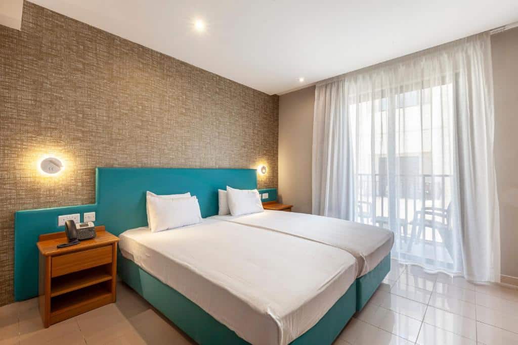 Quarto do Pergola Hotel & Spa com duas camas de solteiro, uma varanda com cadeiras, a cabeceira é azul esverdeado, há duas mesinhas de cabeceira de madeira e iluminação indireta