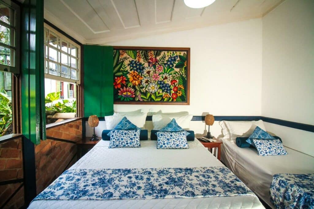 Quarto na Pousada Bartholomeu com uma cama de casal e uma de solteiro, tudo em tons de branco e azul, incluindo as roupas de cama, há uma janela ampla com vista para o jardim e um quadro com flores desenhadas na parede sob a cama
