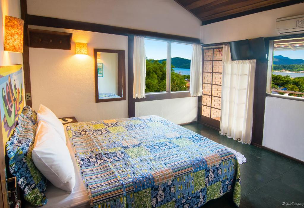 Quarto na Pousada Morro do Forte com as janelas dando vista para o mar, há uma cama de casal, ar-condicionado e um espelho, tudo decorado em tons de branco e com cobertas coloridas sob a cama